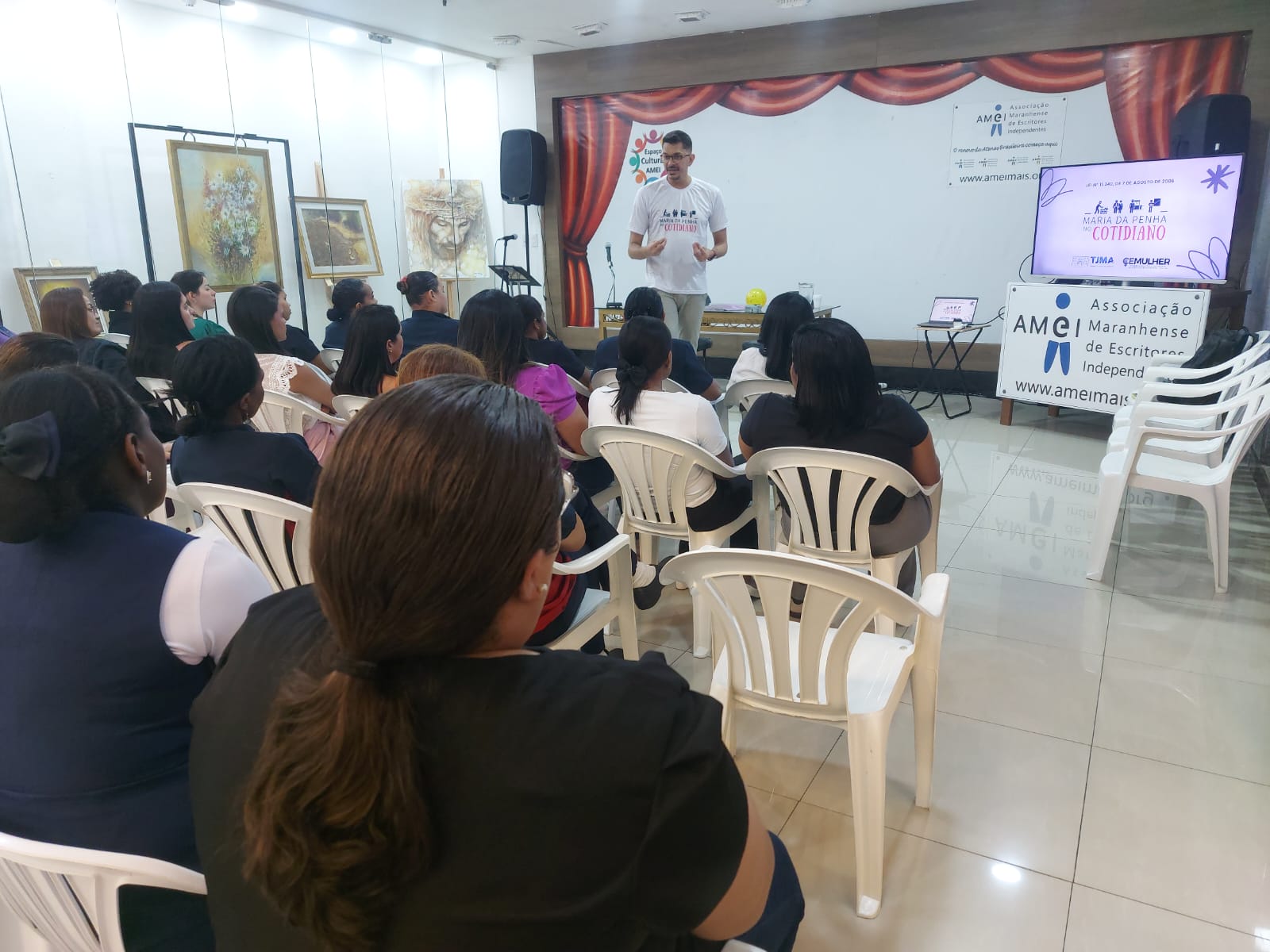 A imagem mostra um evento organizado pela Associação Maranhense de Escritores Independentes (AMCI). Há um grupo de pessoas sentadas em cadeiras de plástico brancas, assistindo a uma apresentação. O apresentador, vestindo uma camiseta branca com texto, está em pé diante do público. Ao lado direito dele, há uma grande tela exibindo informações como o nome da associação e seu site, "ameimais.org". Também há um título destacado, "MARANHENSE DA PENNA COTIDIANO", junto com os nomes "TIMA" e "EMULHER". O ambiente tem quadros decorativos nas paredes, incluindo pinturas de flores e de um leão.