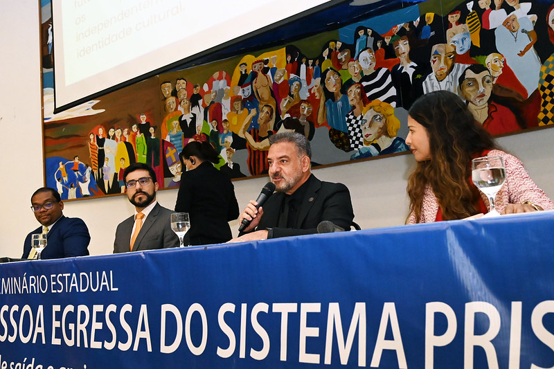 Foto retangular. Três homens e uma mulher estão sentados em bancada. Na bancada, parte do texto em branco: Seminário Estadual de Política de Atenção à Pessoa Egressa do Sistema Prisional, em fundo azul. Ao fundo, pintura colorida de pessoas e tela de projeção.