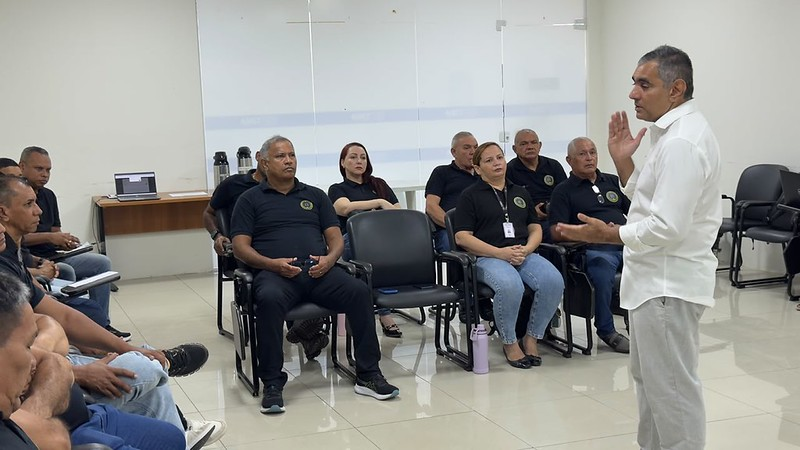 Fotografia em ambiente interno de uma reunião corporativa ou treinamento. À direita, em pé e de perfil, um homem de camisa social branca e calça clara fala enquanto gesticula com as mãos. À sua frente, um grupo de pessoas está sentado em cadeiras de auditório pretas, prestando atenção. A maioria dos ouvintes veste camisas polo pretas com um brasão dourado no peito esquerdo. O ambiente é bem iluminado, com paredes brancas, piso claro e, ao fundo, vê-se uma divisória de vidro e uma mesa com garrafas térmicas.
