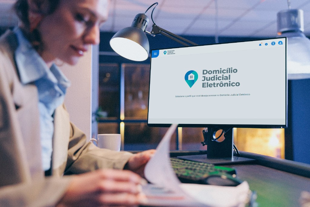 A imagem mostra uma pessoa sentada em uma mesa, olhando para um papel em suas mãos. Na frente da pessoa, há um monitor de computador exibindo uma página da web com o título ¿Domicílio Judicial Eletrônico¿. A página parece estar em português e parece estar relacionada a serviços judiciais eletrônicos. A pessoa está usando óculos e uma camisa azul, e há uma lâmpada na mesa iluminando o espaço de trabalho. O ambiente tem uma iluminação suave.