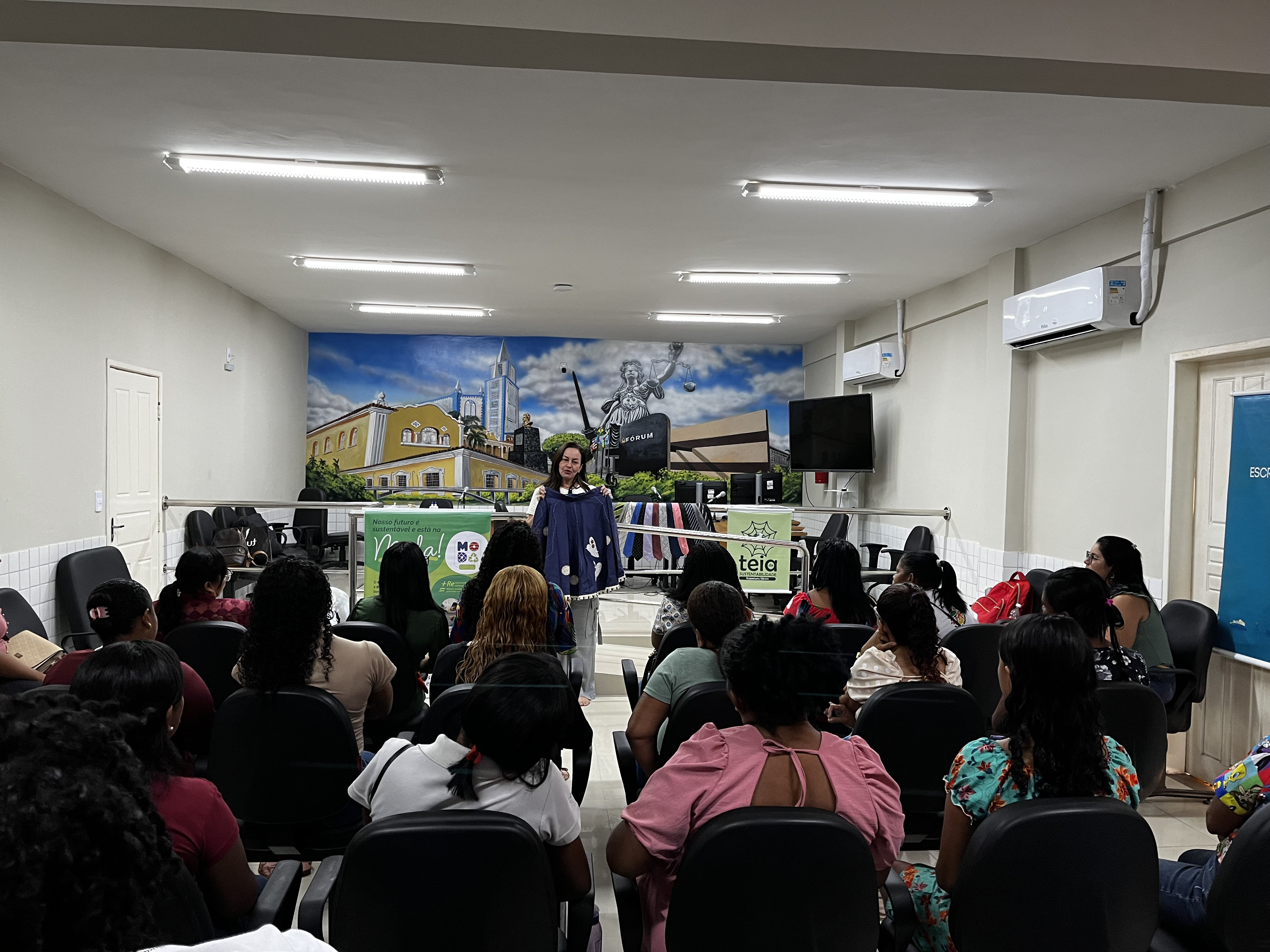 A IMAGEM APRESENTA PLATEIA EM OFICININA. AO FUNDO, A PALESTRANTE APRESENTA UMA SAIA NA COR AZUL AOS PRESENTES NO SALÃO DO JÚRI DE ITAPECURU.
