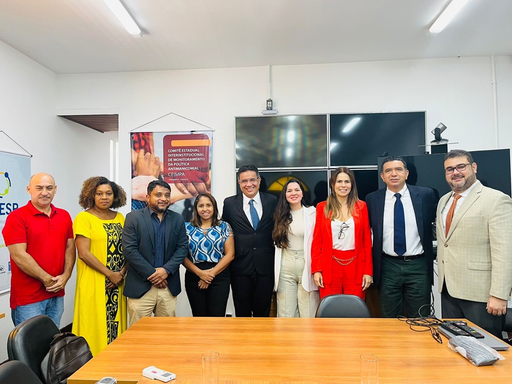 Foto em ambiente interno de um grupo de pessoas vestidas formalmente, em pé, lado a lado, em frente a uma mesa retangular