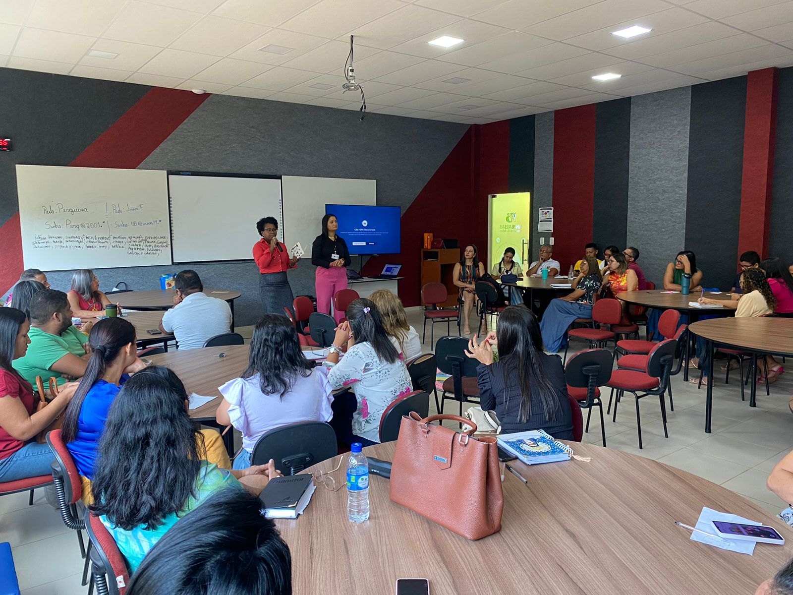 Foto de capacitação. Sala com paredes com listras pretas, cinza e vermelha. Mulheres e homens sentados em mesa. Ao centro, Duas mulheres ao lado de uma televisão.