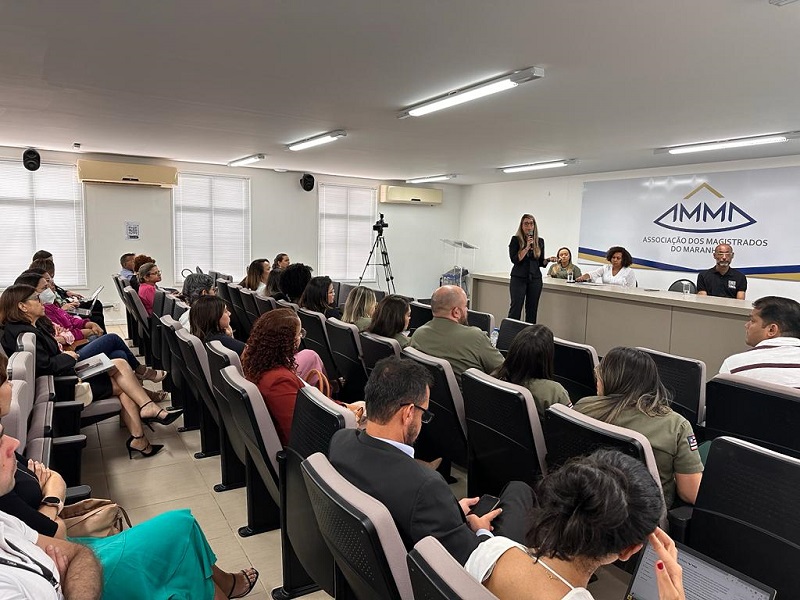 Foto a partir do fundo e á direita, de auditorio com pessoas sentadas, e à frente, de pé, a juíza Mirella Freitas fazendo apresentação.atrás dela, três pessoas estão à mesa de trabalho do seminário