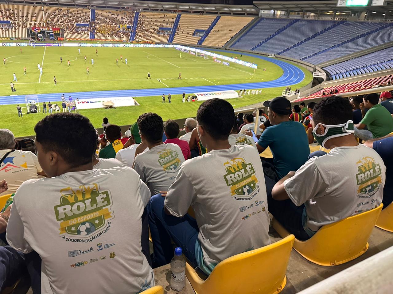 Foto colorida durante a noite, mostrando visão da parte de cima de arquibancadas de estádio, onde abaixo aparecem jovens de costas sentados, usando camisas com logomarca "Rolê do Esporte" nas costas. Abaixo, ao fundo, aparece o lado direito do campo de futebol com jogadores em campo.