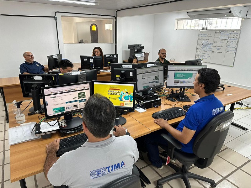 foto em ambiente interno de sala corporativa, com paredes brancas ao fundo. Ao centro, servidores e servidoras trabalham em frente a computadores, estando cinco deles de frente e dois de costa. na parede à direita, um quadro branco com informações manuscritas