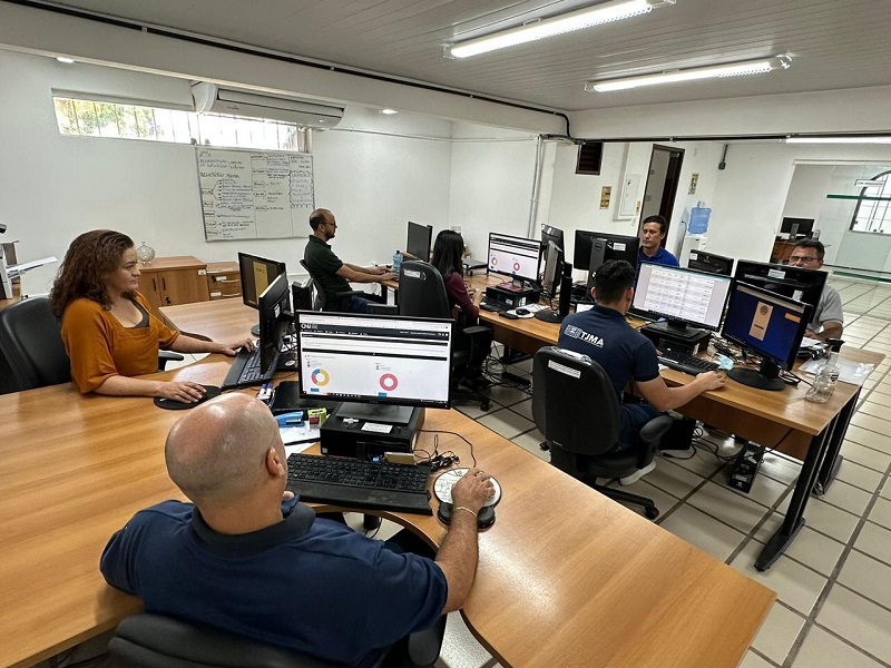 foto em ambiente interno de sala corporativa, com paredes brancas ao fundo. Ao centro, servidores e servidoras trabalham em frente a computadores com sistemas mostrados nas telas. na parede à esquerda, um quadro branco com informações manuscritas