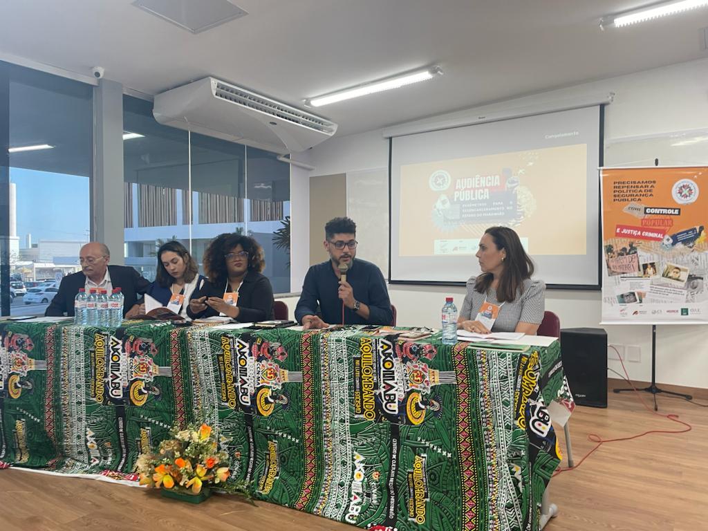 Foto em ambiente interno (sala), à uma mesa coberta com tecidos com estampas africanas, na qual estão autoridades sentadas, entre elas o juiz José dos Santos Costa. O advogado Jorge Serejo está de pé fazendo uso da fala