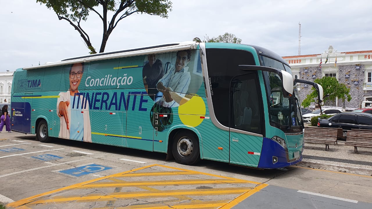 A imagem mostra um ônibus colorido estacionado em uma área externa, com edifícios e árvores ao fundo. O ônibus é azul e verde, com imagens de pessoas e texto impresso em sua lateral. O texto ¿Conciliação Itinerante¿ está visível na lateral do ônibus, indicando um serviço móvel de conciliação. Há uma imagem de uma pessoa com a mão estendida, sugerindo um aperto de mão. O ônibus está estacionado em uma área marcada com linhas amarelas no chão. Ao fundo, há edifícios brancos e árvores, indicando que o ônibus está em um ambiente urbano. O céu é parcialmente nublado.