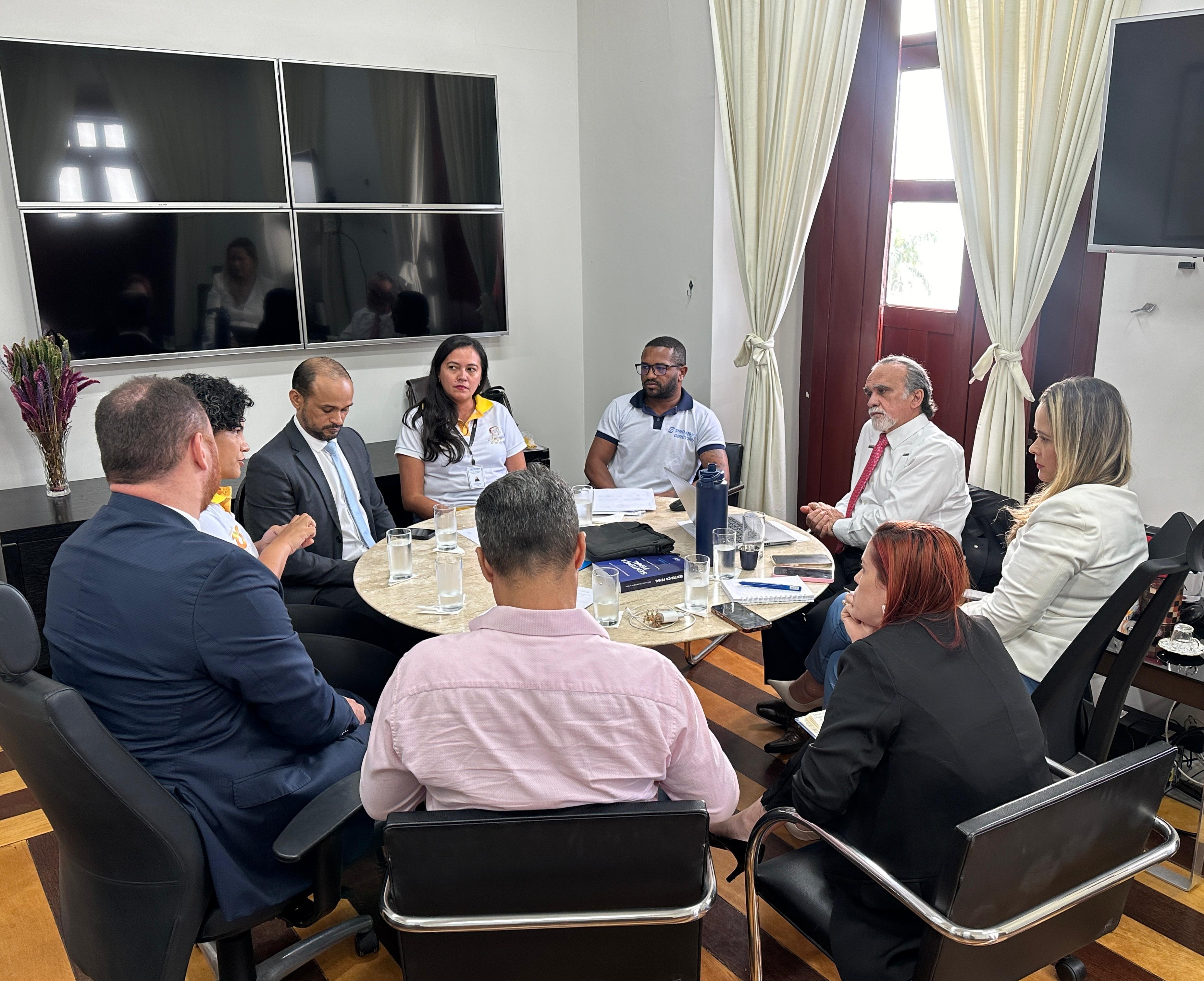 A imagem retrata um grupo de pessoas sentadas ao redor de uma mesa de vidro em um ambiente que parece ser uma reunião formal. Os rostos das pessoas foram obscurecidos com blocos de cor para preservar a privacidade. Há um buquê de flores na mesa, e a sala possui cortinas e vários monitores na parede.