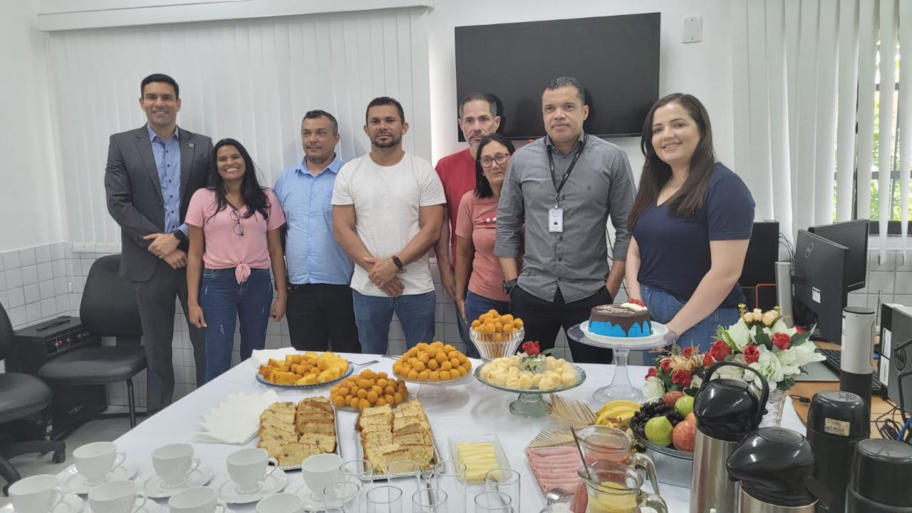 imagem mostra um grupo de pessoas reunidas em torno de uma mesa em um ambiente interno, provavelmente para uma reunião ou celebração.
