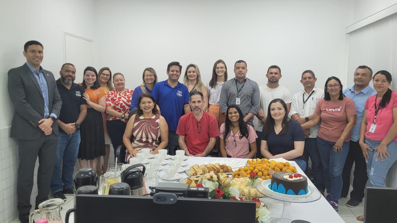 imagem mostra um grupo de pessoas reunidas em torno de uma mesa em um ambiente interno, provavelmente para uma reunião ou celebração.