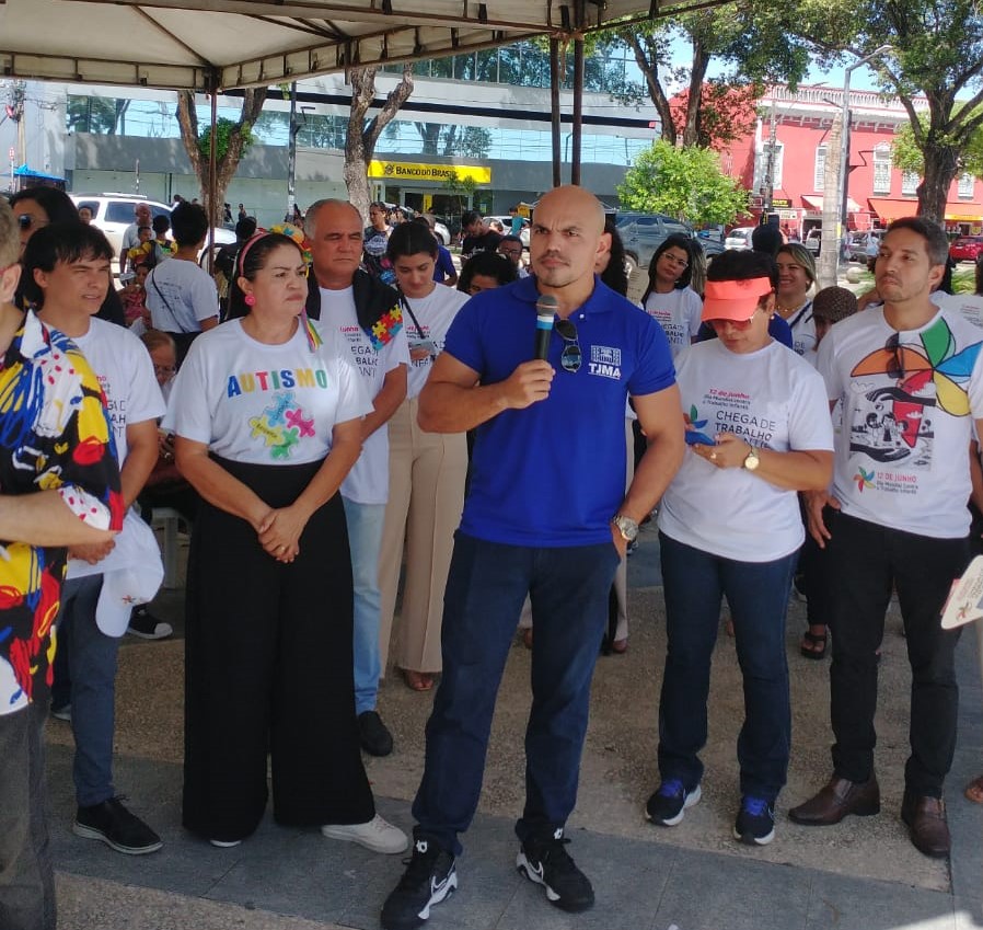 A imagem mostra um grupo de pessoas reunidas sob uma tenda branca.  Elas estão vestindo roupas variadas, incluindo camisetas com diferentes estampas e cores, sugerindo um evento casual ou encontro. Alguns indivíduos seguram papéis ou pequenos objetos nas mãos.