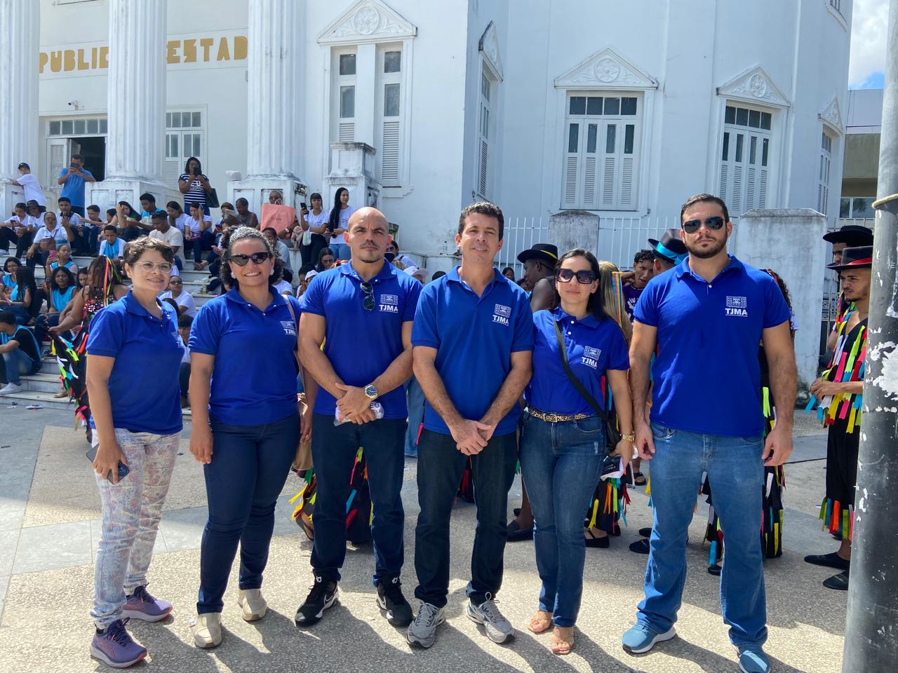 A imagem mostra um grupo de sete pessoas em frente a um prédio com a placa ¿BIBLIOTECA PÚBLICA¿ acima da entrada. As pessoas estão vestindo camisas azuis com logotipos visíveis, possivelmente indicando que fazem parte de uma organização ou evento.  Ao fundo, há uma multidão de pessoas sentadas e em pé, sugerindo que algum evento público está acontecendo