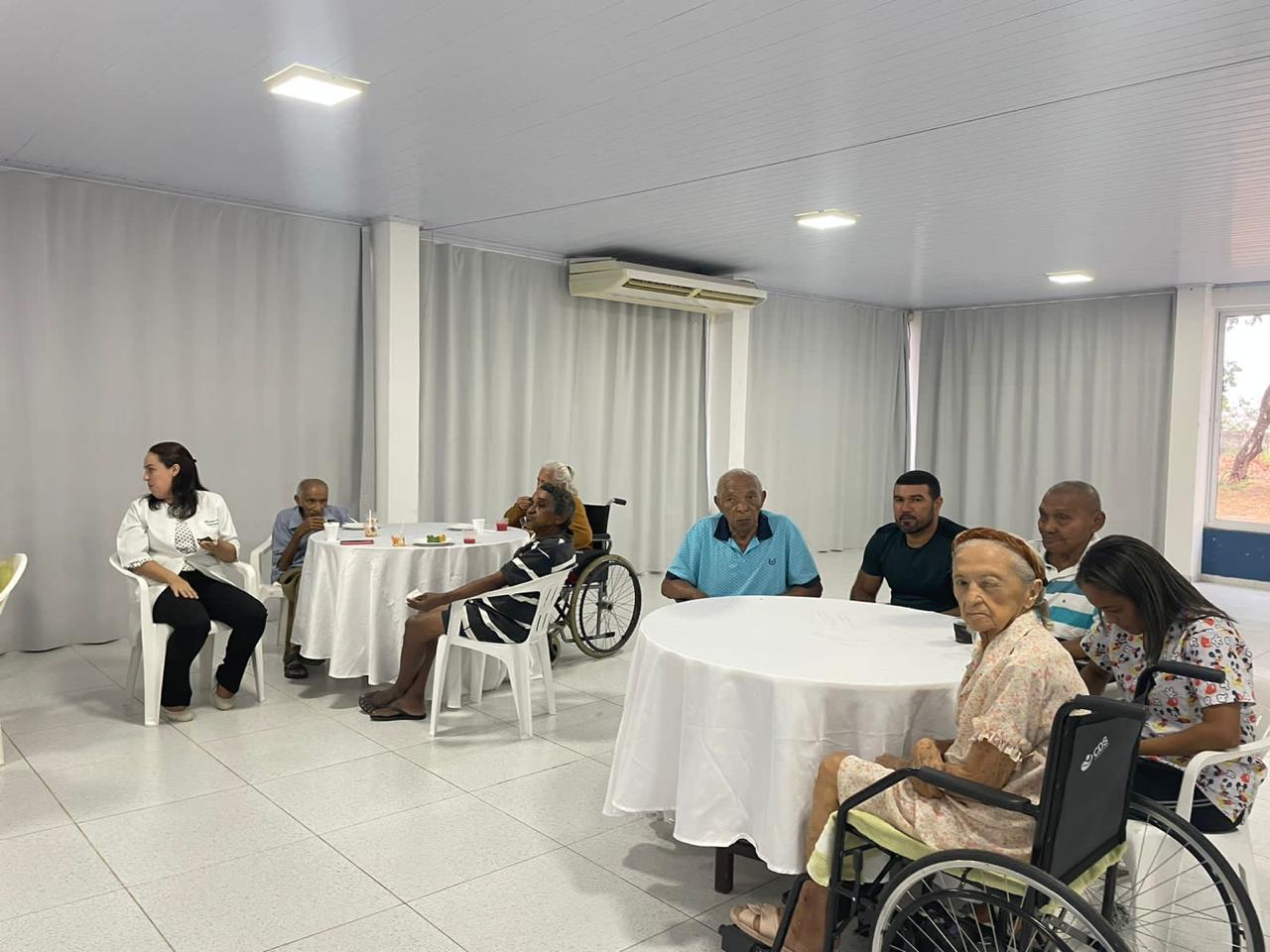 Essa imagem mostra um ambiente interno com várias pessoas sentadas ao redor de duas mesas redondas cobertas por toalhas brancas. São visíveis sete indivíduos, alguns de frente para a câmera e outros de costas. Um deles está em uma cadeira de rodas. A sala tem paredes e cortinas brancas, e há um aparelho de ar-condicionado. Há pratos, copos e o que parece ser uma garrafa nas mesas, sugerindo um evento social ou refeição. A iluminação no ambiente é clara.