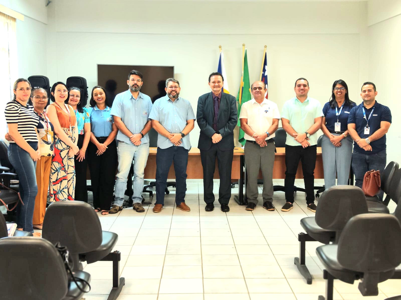 A imagem mostra um grupo de onze pessoas em uma sala com cadeiras dispostas em fileiras, voltadas para a câmera, sugerindo um ambiente formal ou profissional. As pessoas estão posicionadas em uma linha Ao fundo, há bandeiras em mastros, incluindo o que parece ser a bandeira do Brasil com base em suas cores e design. Essa imagem pode ser relevante ou interessante devido ao contexto da reunião ou evento em andamento, indicado pela disposição formal e trajes das pessoas, bem como pela presença das bandeiras nacionais, que sugerem uma natureza oficial ou governamental.