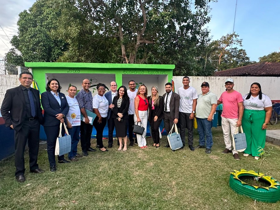 A imagem mostra um grupo de catorze indivíduos ao ar livre, em frente a uma estrutura verde com telhado branco. A maioria segura o que parece serem certificados ou documentos, sugerindo algum tipo de cerimônia ou evento oficial. Há também um objeto no chão, na frente, que se assemelha a uma mangueira ou cabo enrolado, próximo a uma pequena estrutura verde, possivelmente relacionada a jardinagem ou manutenção. O cenário parece ser um parque ou área recreativa devido à presença de grama e árvores ao fundo. NAa parede ao fundo, o texto "Ecoponto"