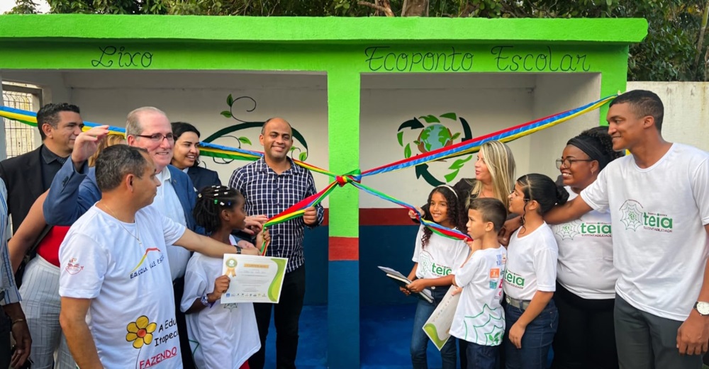 Grupo de adultos e crianças partiicipa de cerimonia de corte de fita em ambiente externo. As crianças vestem camisas brancas com marca da "Teia da Sustentabilidade"