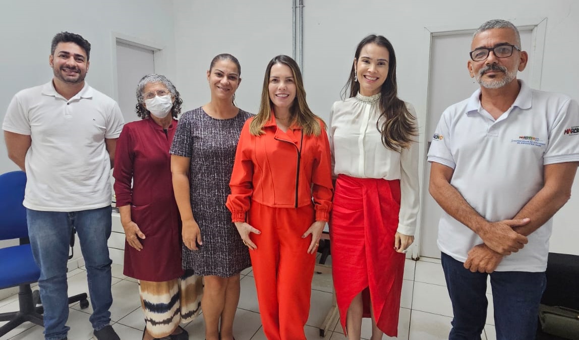 FOTO HORIZONTAL, COLORIDA, DE HOMENS E MULHERES EM TRAJES FORMAIS, DE PÉ, POSANDO PRA FOTO EM SALA DE PAREDES BRANCAS.