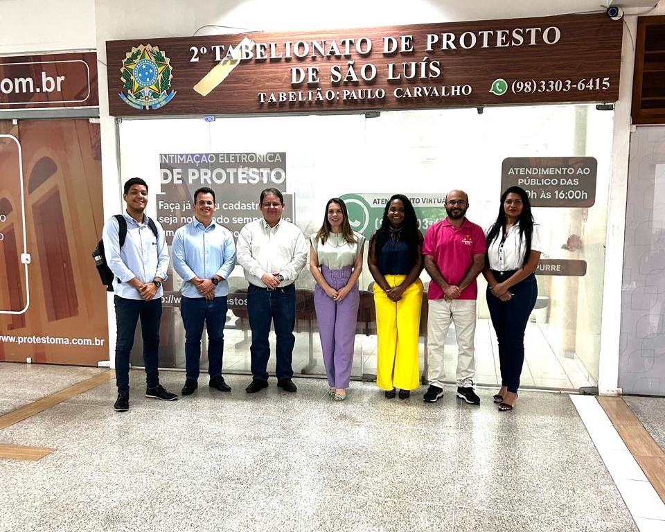 Foto horizontal, colorida, de seis pessoas em frente a uma fachada de cartório Na parede atrás do grupo, há placas e informações. Uma delas diz "2º TABELIONATO DE PROTESTO DE SÃO LUÍS,". Outra placa menor m À direita, há uma placa indicando o horário de atendimento ao público das 8:00 às 16:00 e mencionando "TABELIÃO PAULO CARVALHO".