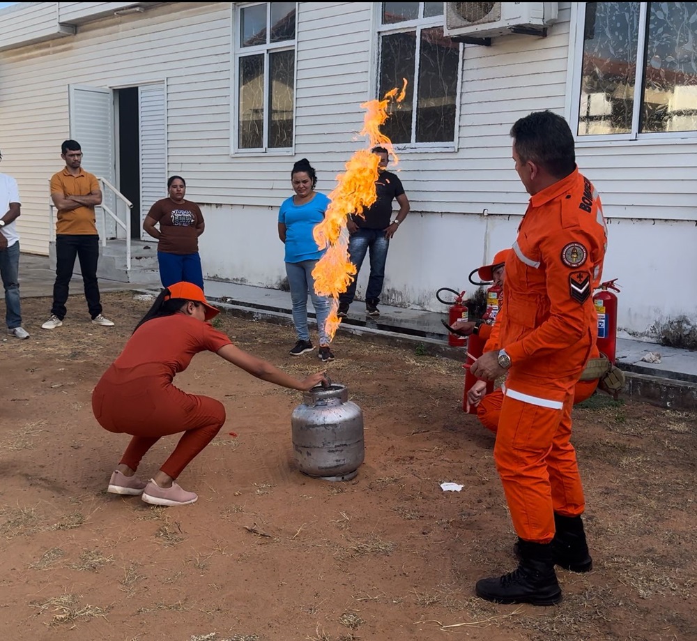 Em ambiente externo, mulher agachada manipula botijao de gás em chamas ao lado de agente do Corpo de Bombeiros e outras pessoas observam