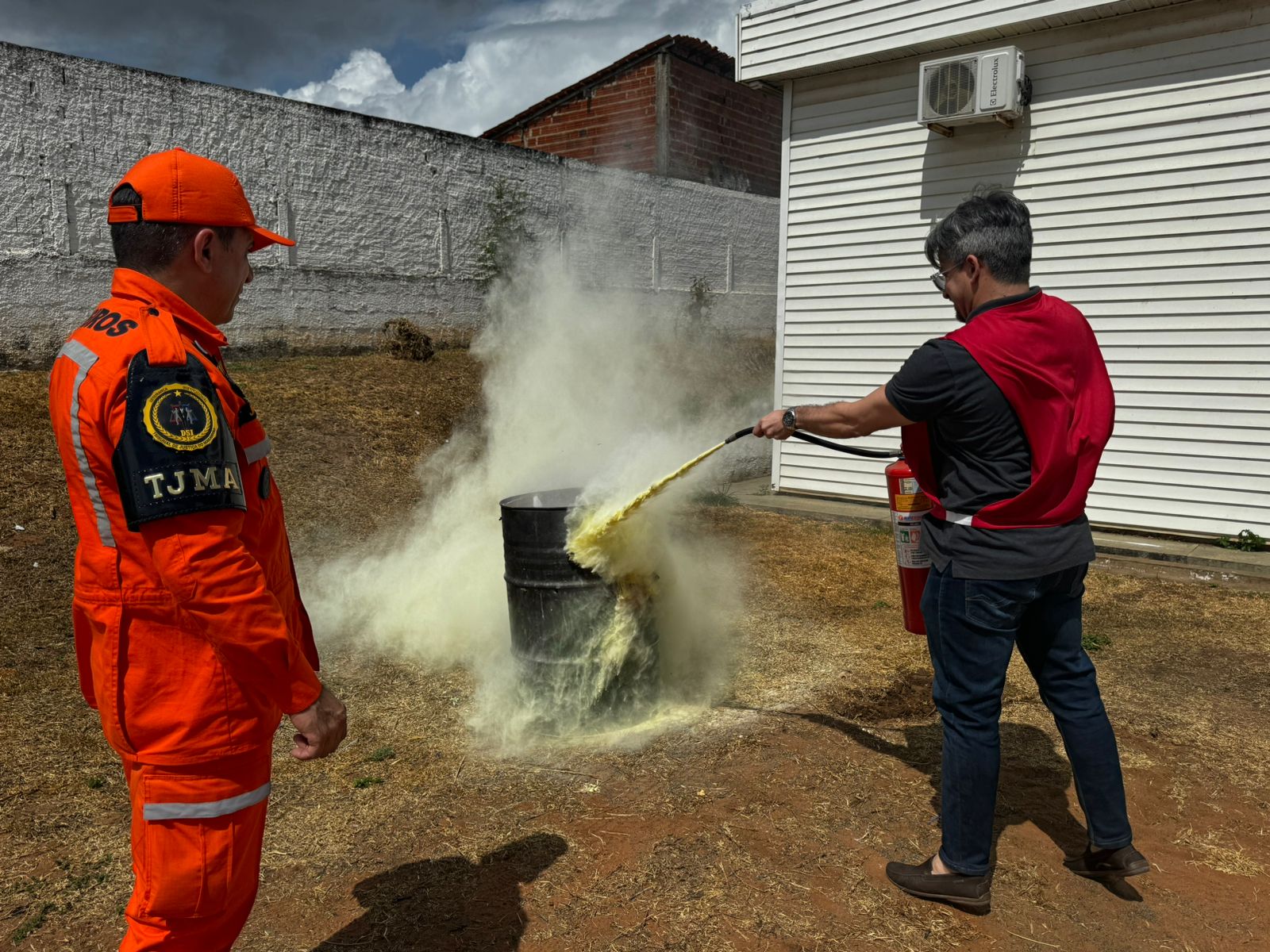 A imagem retrata duas pessoas ao ar livre. Uma delas está vestindo um uniforme laranja com as iniciais ¿TJMA¿, enquanto a outra está com roupas casuais. A pessoa de roupas casuais segura uma mangueira, borrifando uma substância branca em um barril preto que emite fumaça. O cenário parece ser um quintal com uma parede branca e estruturas de tijolos vermelhos ao fundo.