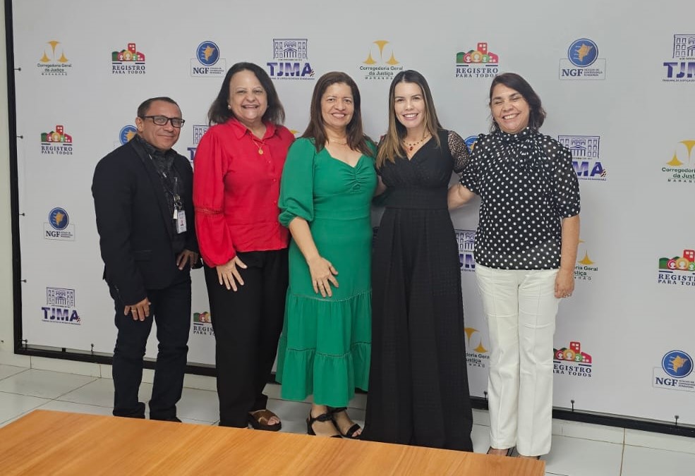 Foto horizontal, colorida, de quatro mulheres e um homem de pé, lado a lado, posando pra foto, vestindo roupa formal. Eles estão em frente a um fundo branco com vários logotipos de instituições.