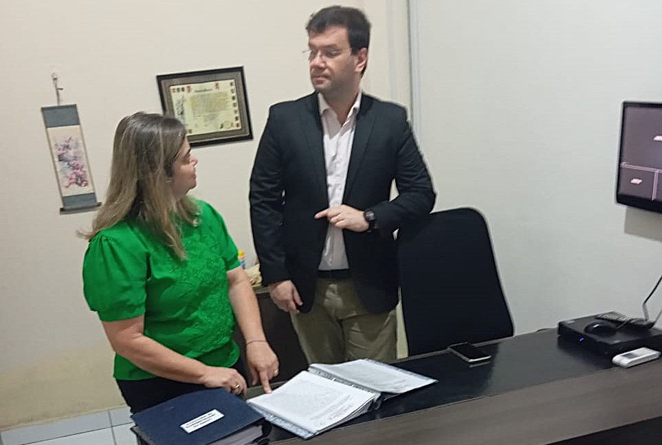Foto horizontal, colorida, mostrando duas pessoas em um escritório. Um homem e uma mulher conversam de pé atrás de mesa. A mulher à esquerda está vestindo uma camisa verde com mangas curtas O homem usa um blazer preto sobre uma camisa branca com a gola aberta e calças cáqui. No fundo, há um monitor de computador sobre um móvel e quadros decorativos na parede.