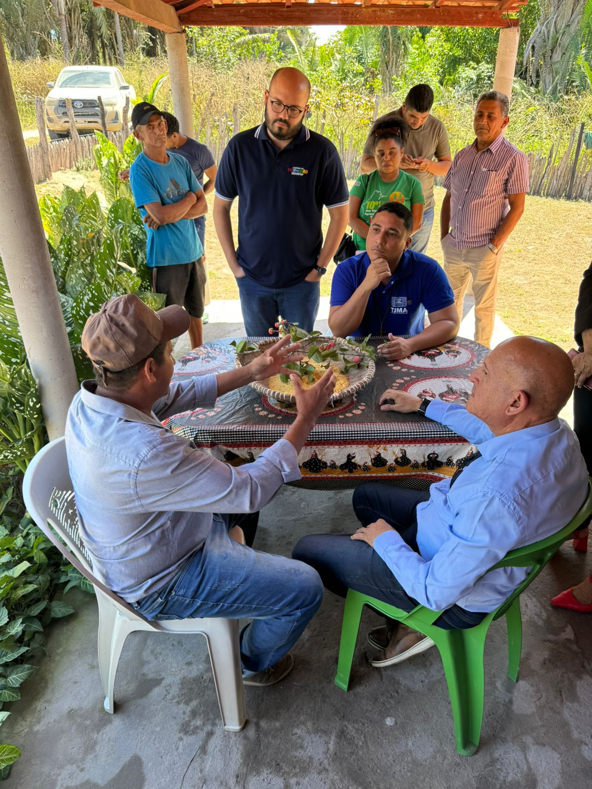 A imagem retrata um grupo de pessoas reunidas ao redor de uma mesa, com os rostos obscurecidos para preservar a privacidade. O cenário parece ser ao ar livre, sob um abrigo com telhado, e há um veículo visível ao fundo. Na mesa, há o que parece ser comida sendo compartilhada ou servida. As pessoas estão envolvidas em uma atividade conjunta, o que pode indicar um evento social ou encontro.