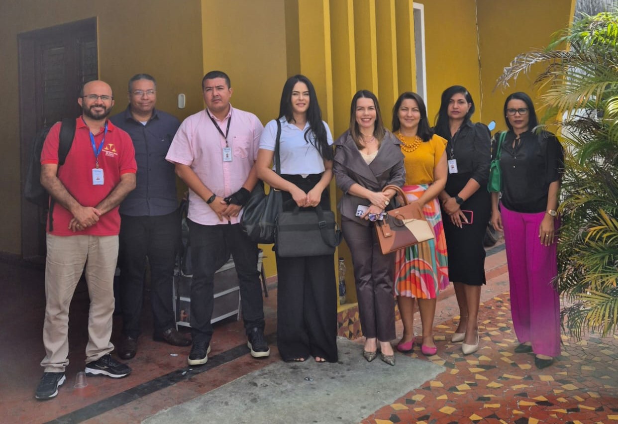 Foto horizontal, colorida, mostra um grupo de onze pessoas, entre homens e mulheres, de pé em frente a um prédio com pilares amarelos. O grupo posa pra foto, olhando pra frente; alguns usando roupas formais e outros vestidos de maneira mais casual.