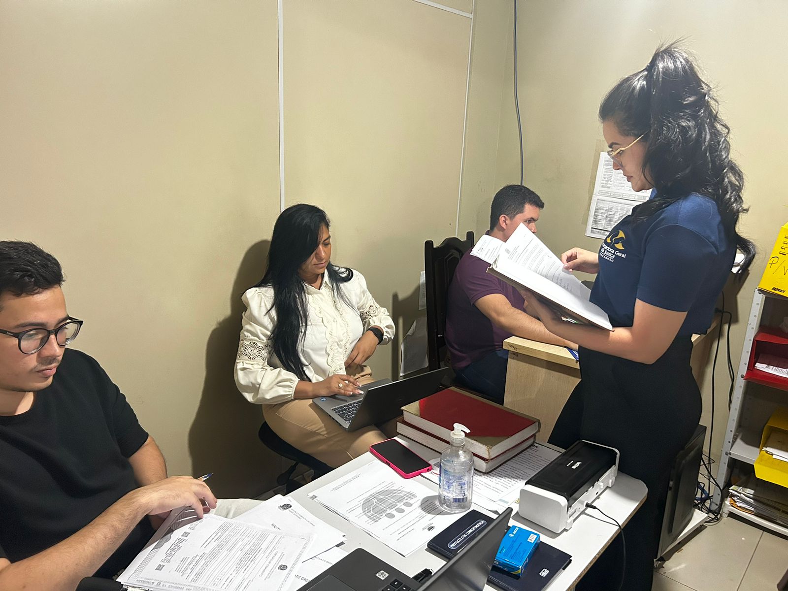 Foto horizontal, colorida, mostrando quatro pessoas em escritório. Três estão sentados em mesas com computadores e papéis, enquanto a quarta está de pé segurando alguns papéis. Há uma parede bege ao fundo.