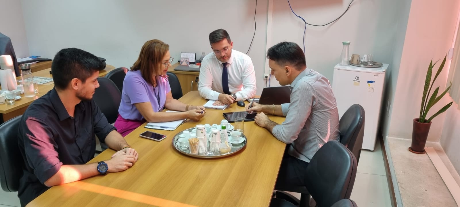 A imagem mostra quatro pessoas sentadas ao redor de uma mesa retangular em um ambiente de escritório. Seus rostos estão obscurecidos por retângulos marrons para preservar a privacidade. A pessoa à esquerda usa um relógio escuro e tem os braços cruzados. As duas pessoas no meio parecem estar envolvidas em uma discussão, com uma delas segurando uma caneta e apontando para um documento na mesa, sugerindo que podem estar analisando ou discutindo seu conteúdo. A pessoa à direita está usando um laptop. Na mesa, há vários itens, incluindo documentos, telefones celulares, garrafas de água e óculos empilhados ordenadamente. Ao fundo, há um bebedouro de escritório e o que parece ser uma janela com persianas parcialmente fechadas.