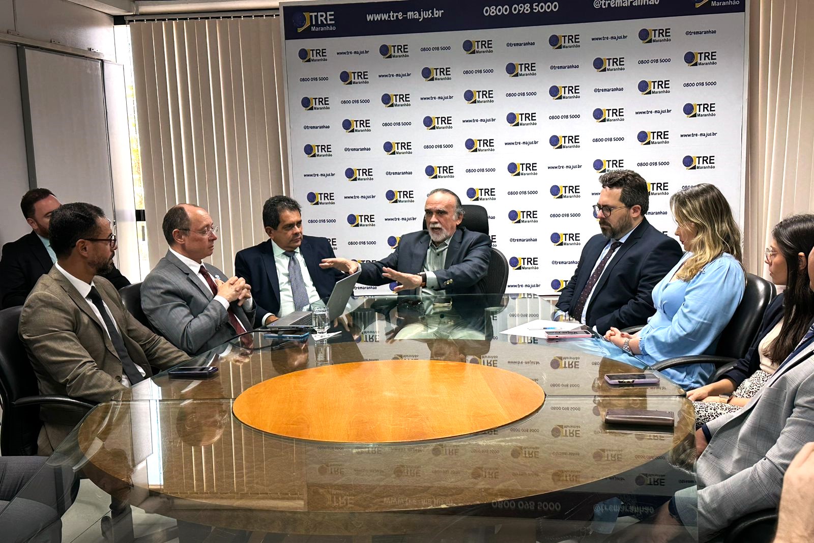 A imagem mostra um grupo de pessoas sentadas ao redor de uma mesa de conferência. O cenário é de uma reunião formal. Ao fundo, há uma faixa com a inscrição ¿TRE¿.