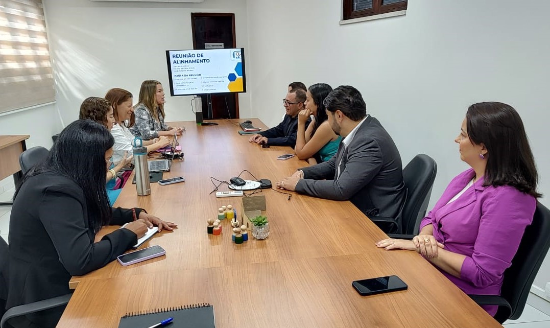 Foto horizontal, colorida, de homens e mulheres sentadas ao redor de uma mesa em uma sala de reuniões. Há uma tela de apresentação na frente da sala exibindo conteúdo. Algumas pessoas têm cadernos e canetas sobre a mesa.