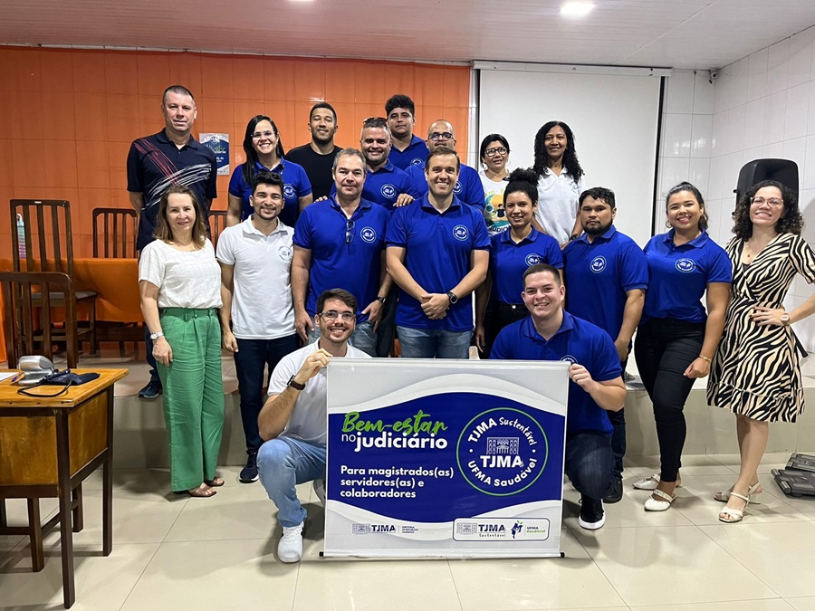 Imagem em ambiente interno com um grupo de pessoas, a maioria vestindo camisas azuis iguais, posam para foto exibindo banner do projeto "Bem estar no Judiciário"