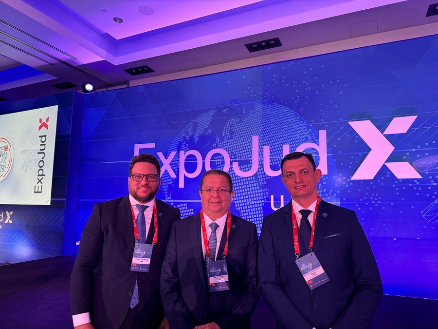 Os magistrados Felipe Damous, Jamil Gedeon e Rodrigo Terças, do TJMA, posando para foto em ambiente interno~, vestindo ternos e usando crachás do evento. Atrás deles, fundo de palco da Expojud X USA