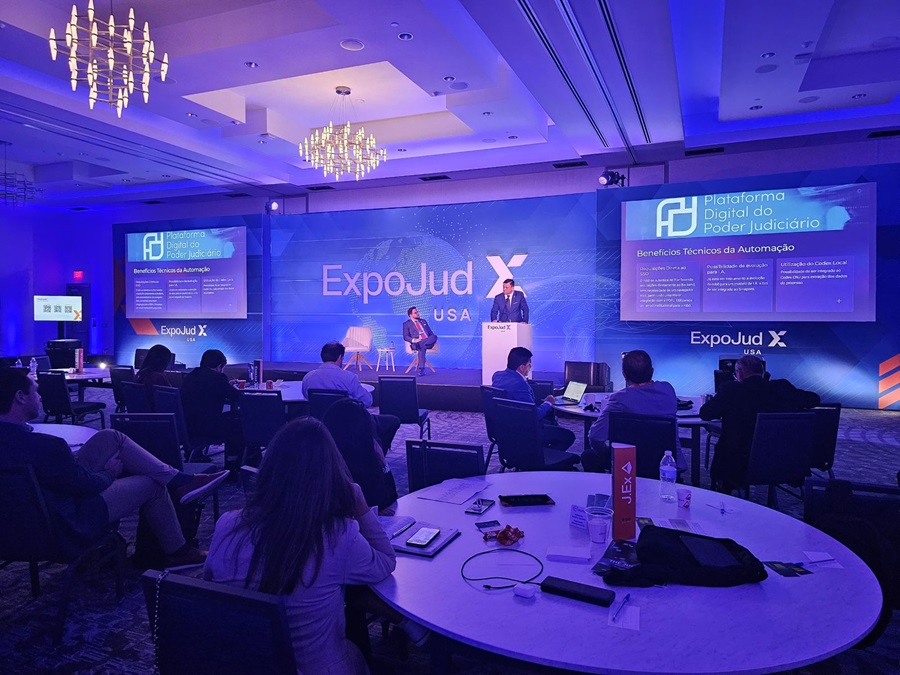 A imagem mostra um evento indoor com o juiz Rodrigo Terças como palestrante apresentando no palco, na frente de uma sala. O público está sentado em mesas redondas, e há laptops abertos, sugerindo que os participantes podem estar tomando notas ou acompanhando a apresentação. A sala tem carpete azul e lustres modernos pendurados no teto. No palco, há uma grande tela exibindo um slide de uma apresentação com gráficos e texto relacionados ao ¿ExpoJud¿ e ¿Plataforma Digital do Judiciário.¿ O slide menciona ¿Interfaces: Sociedade de Automação.¿ Há banners em ambos os lados do palco com logotipos do ExpoJud USA.