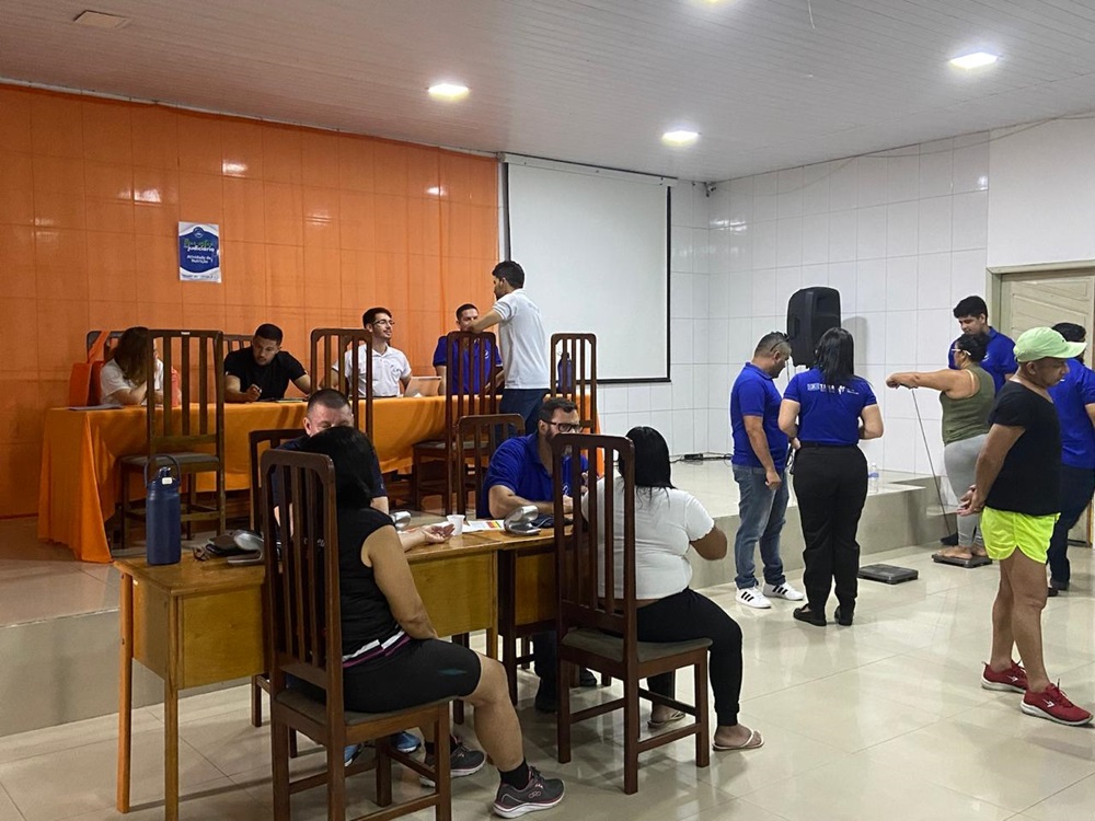 A imagem retrata um ambiente interno com um grupo de pessoas. Algumas estão sentadas em mesas, enquanto outras estão em pé. Na frente, há duas mesas dispostas em formato de T, com cadeiras ao redor. No fundo, um grupo de pessoas está em pé, interagindo próximo a uma área que parece ser um balcão com banquetas. As paredes são pintadas de laranja, e há uma faixa pendurada com texto e possivelmente um logotipo. Alto-falantes estão fixados no teto. Todos na imagem parecem estar envolvidos em conversas ou atividades