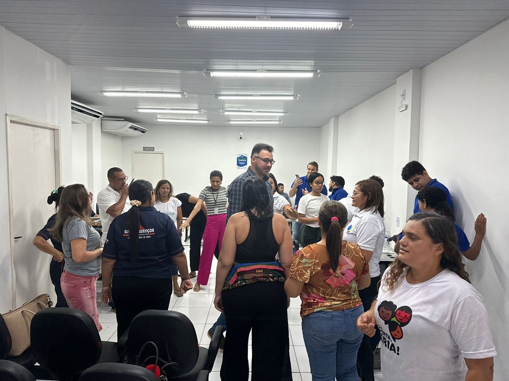 A imagem retrata um ambiente interno, provavelmente uma sala de aula ou sala de treinamento, com paredes brancas e iluminação fluorescente. Várias pessoas estão em pé, voltadas para a frente da sala, onde alguém parece estar conduzindo uma sessão ou apresentação. Há cadeiras pretas dispostas em fileiras, algumas ocupadas e outras vazias. Um quadro branco é parcialmente visível na frente da sala, mas nenhum texto pode ser discernido dessa perspectiva. A cena sugere uma atividade em grupo ou ambiente de aprendizado onde a interação está ocorrendo.