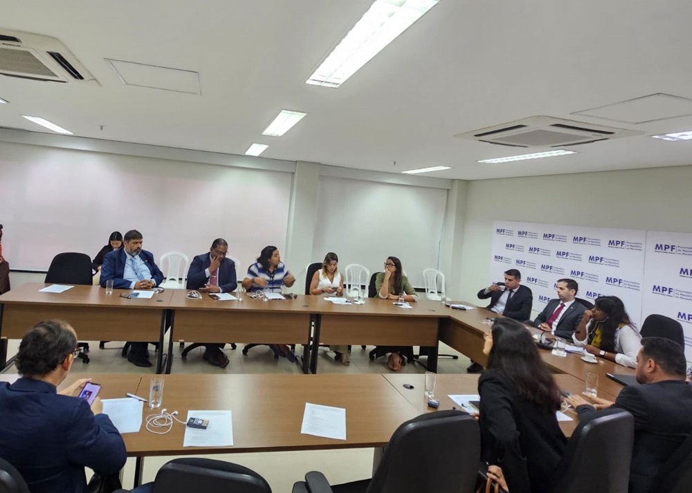 A imagem mostra uma reunião formal acontecendo em uma sala de conferências. Há várias pessoas sentadas ao redor de uma grande mesa retangular. Alguns participantes estão de frente para a câmera, enquanto outros estão de costas. A sala tem paredes brancas e está equipada com móveis modernos, incluindo cadeiras de escritório pretas e a mesa mencionada. No fundo da sala, há um banner com logotipos e texto que parece estar relacionado a instituições ou organizações oficiais, mas os detalhes específicos do texto não são claros à distância. O ambiente sugere uma discussão ou negociação profissional, possivelmente em um contexto corporativo ou governamental.