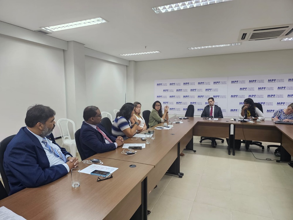 A imagem mostra uma reunião formal acontecendo em uma sala de conferências. Há várias pessoas sentadas ao redor de uma grande mesa retangular. Alguns participantes estão de frente para a câmera, enquanto outros estão de costas. A sala tem paredes brancas e está equipada com móveis modernos, incluindo cadeiras de escritório pretas e a mesa mencionada. No fundo da sala, há um banner com logotipos e texto que parece estar relacionado a instituições ou organizações oficiais, mas os detalhes específicos do texto não são claros à distância. O ambiente sugere uma discussão ou negociação profissional, possivelmente em um contexto corporativo ou governamental.