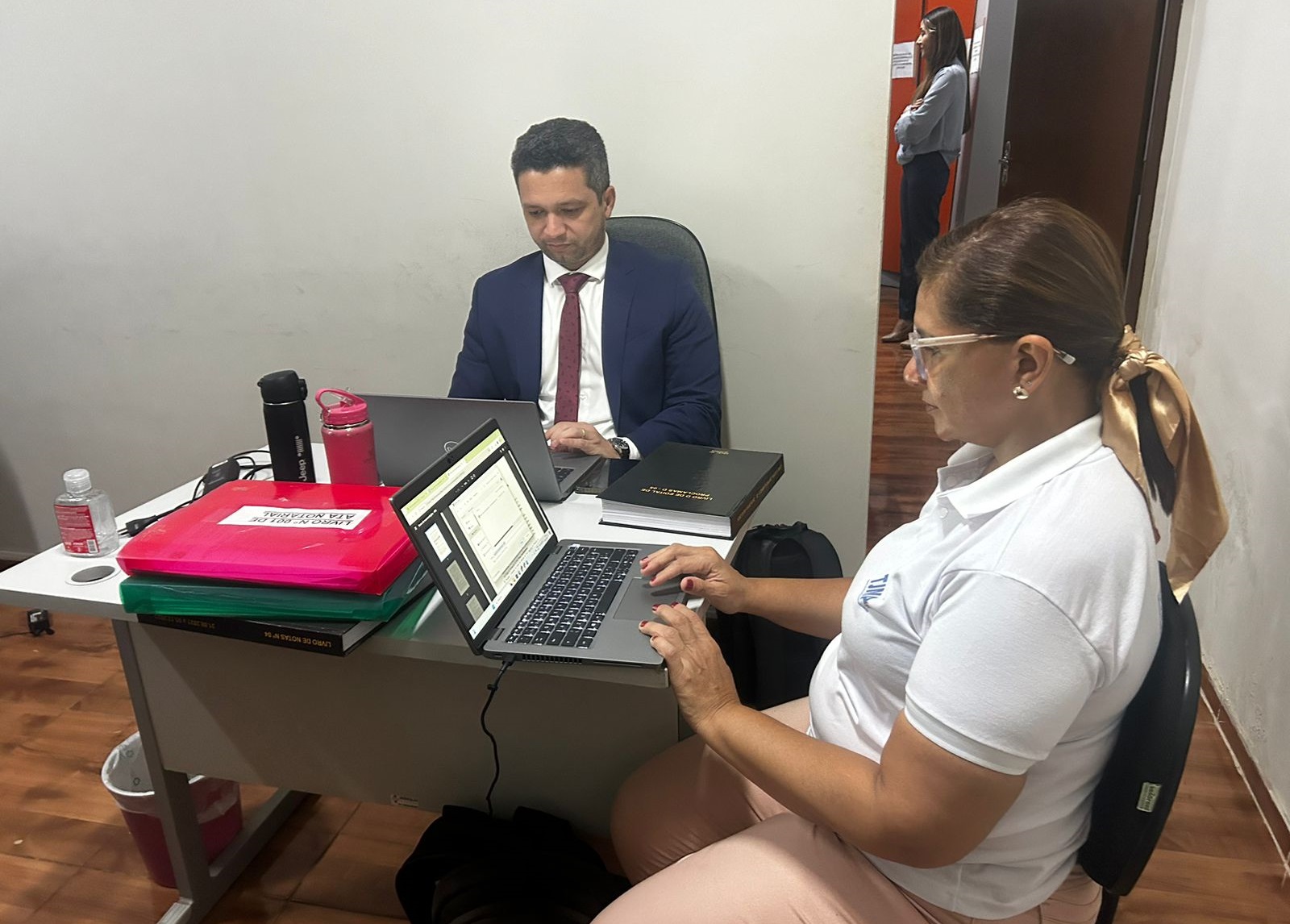 Foto horizontal, colorida, de um homem pardo, de terno escuro e uma mulher branca, cabelos louros, presos, de óculos e camisa branca e calça bege, em um ambiente de escritório. O homem à esquerda está vestindo um terno escuro e está voltado para a tela de um laptop. A mulher, à direita, também está usando outro laptop. Na mesa, há vários itens, incluindo cadernos ou pastas empilhadas em rosa e verde, uma garrafa de água preta e o que parece ser um smartphone. Ao fundo, há uma parede branca com alguns itens pendurados e parte de uma peça de roupa laranja visível no lado direito.