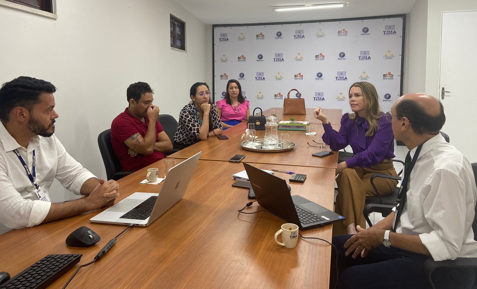 A fotografia mostra um grupo de seis pessoas sentadas ao redor de uma mesa de madeira em uma sala. Ao fundo, cartaz de fundo bvranco com vários logotipos.  Na mesa, há laptops, papéis e copos, sugerindo uma reunião de negócios ou organizacional. Também há um bule transparente com xícaras.