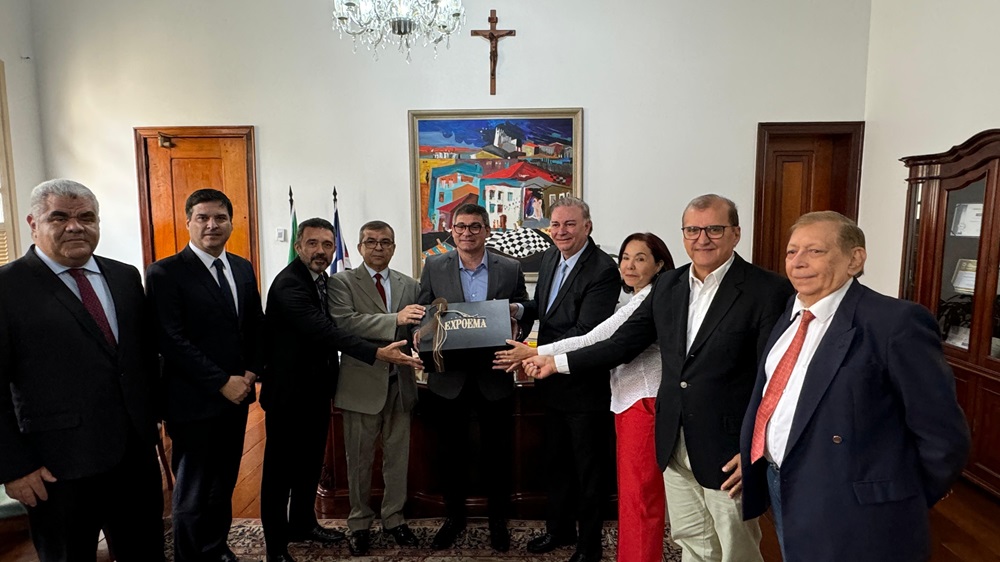 foto em ambiente interno. Grupo de homens em pé, lado a lado, vestindo roupas formais, e exibindo uma caixa preta com a logomarca da Expoema. A sala possui características arquitetônicas tradicionais, como tetos altos e grandes janelas com molduras de madeira.