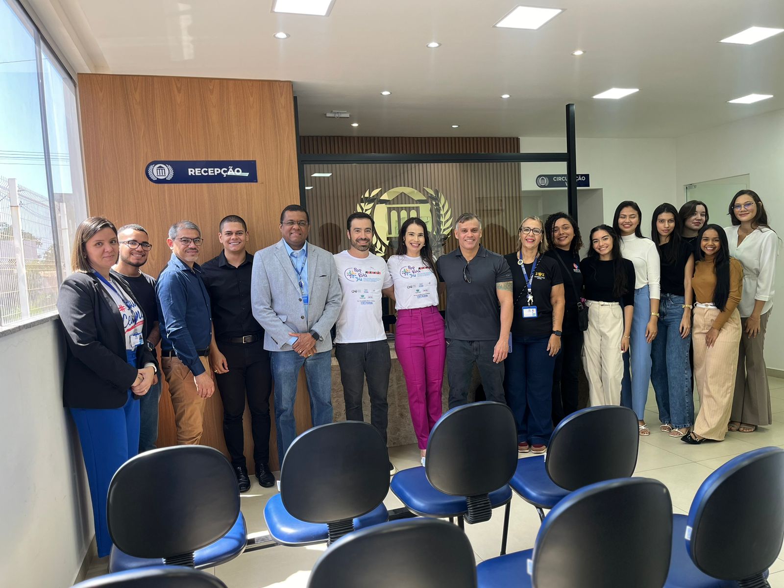 A imagem mostra um grupo de 15 pessoas posando para uma foto em um ambiente fechado. No fundo, há uma parede de madeira clara com um logo institucional no centro, acima de uma porta de vidro, com as palavras "RECEPÇÃO" em destaque em uma placa azul. O chão é de piso claro, e há várias cadeiras pretas com encosto azul organizadas na frente do grupo.

As pessoas estão organizadas em duas fileiras, lado a lado. A maioria usa roupas formais ou semiformais, como blusas, camisas sociais, calças e blazers. Há um misto de homens e mulheres no grupo, todos sorrindo para a câmera. Algumas pessoas usam crachás, indicando que fazem parte de uma organização ou evento. Uma das pessoas está vestindo uma camiseta branca com o logo do evento "PopRuaJud". 

O ambiente é bem iluminado, com luz natural entrando pela janela à esquerda. Ao fundo, é possível ver mais detalhes da sala, que aparenta ser moderna e bem organizada.
