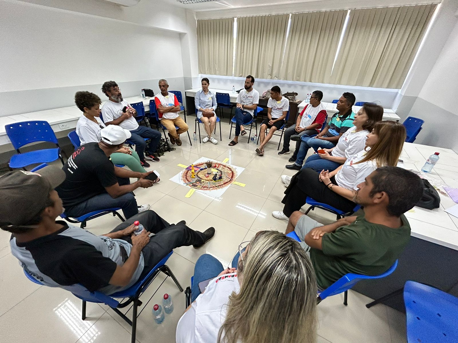 A imagem mostra um grupo de pessoas sentadas em círculo dentro de uma sala. Elas estão participando de uma atividade coletiva, provavelmente uma roda de conversa ou dinâmica de grupo. O chão é claro e o ambiente tem cadeiras azuis espalhadas. No centro do círculo, há um arranjo decorativo composto por elementos coloridos organizados em uma espécie de mandala ou altar, sugerindo uma atividade simbólica ou espiritual.

Os participantes, em sua maioria vestindo camisetas brancas com logos (possivelmente do evento PopRuaJud), estão atentos e envolvidos na atividade. Alguns seguram garrafas de água e outros parecem estar ouvindo alguém que está falando. As janelas ao fundo estão cobertas por cortinas claras, e o ambiente é iluminado por luz artificial.

A sala é simples, com cadeiras de plástico e mesas ao fundo, e parece ser parte de uma instituição ou centro comunitário. As pessoas no círculo estão distribuídas de forma igualitária, criando um clima de interação e diálogo entre todos os presentes.