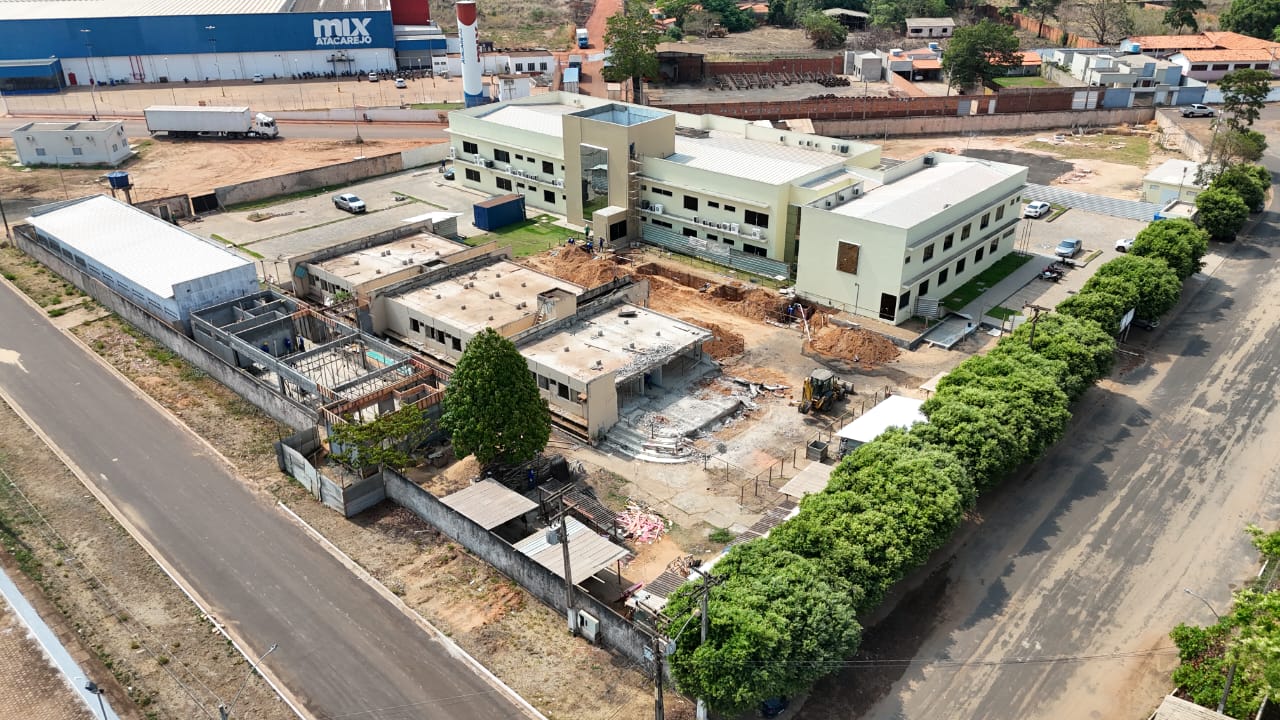 A imagem mostra uma visão aérea de um canteiro de obras. O local inclui vários edifícios, alguns dos quais parecem estar em construção ou renovação. Há um grande prédio concluído com fachada de cor clara e várias unidades de ar-condicionado instaladas no exterior. Ao lado deste edifício, há várias estruturas menores em diferentes estágios de construção, com armações expostas e materiais de construção visíveis. A área é cercada por uma estrada pavimentada e uma linha de árvores de um dos lados. No fundo, há um grande armazém ou edifício industrial com o texto "mix ATACAREJO" escrito. O local também inclui veículos estacionados e equipamentos de construção.