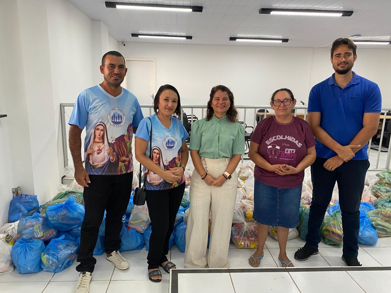 Foto horizontal, colorida, de cinco pessoas em pé em salão de júri com paredes e piso brancos, diante de um gradil branco. Atrás delas, há várias sacolas cheias de itens variados, possivelmente doações ou suprimentos. Duas das pessoas vestem camisas azuis com a imagem de um símbolo religioso e o texto "ESCOLHIDOS POR DEUS.
