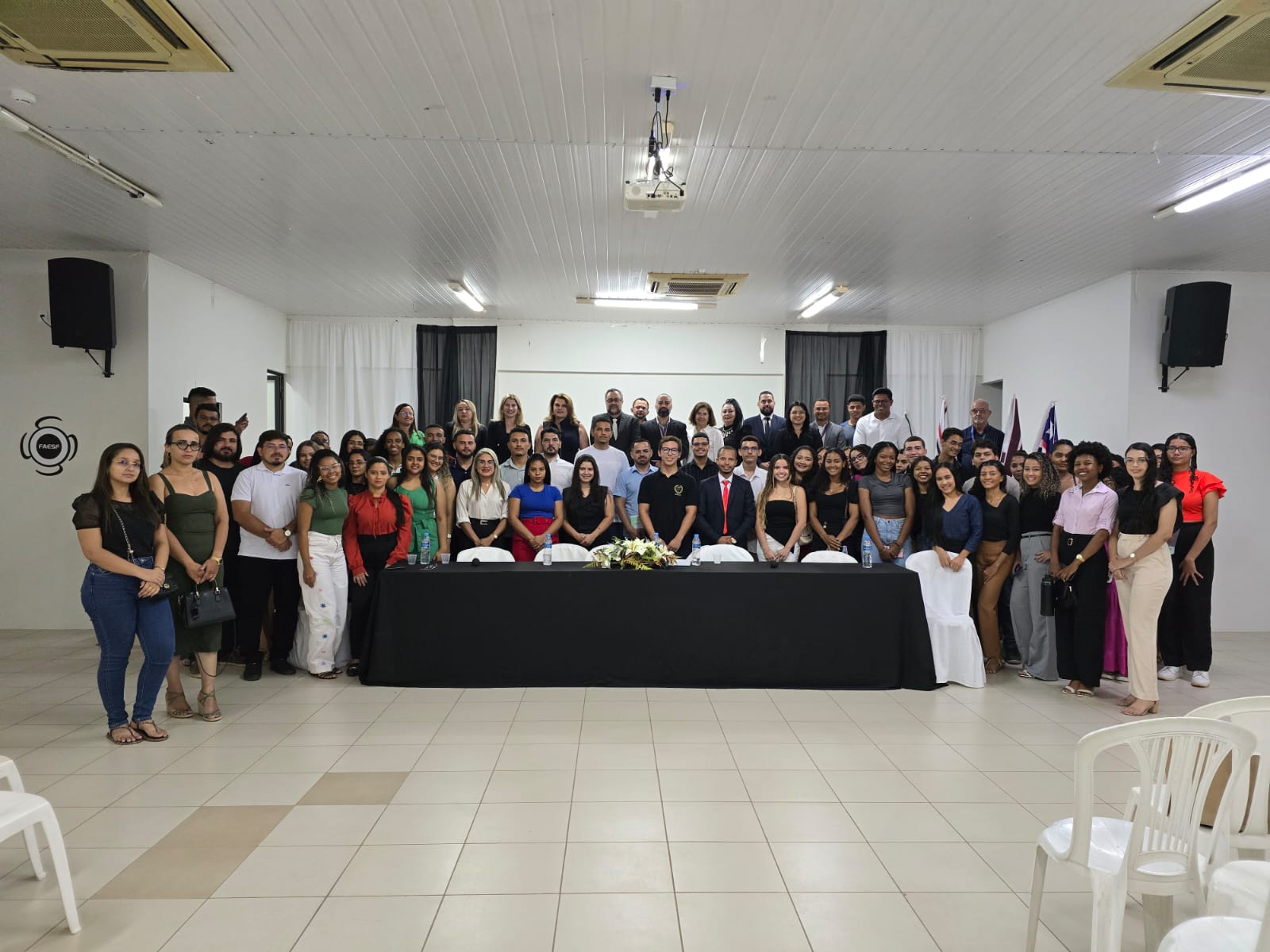 A imagem mostra um grupo grande de pessoas posando para uma foto em um ambiente interno. As pessoas estão organizadas em várias fileiras, com algumas sentadas atrás de uma mesa coberta com uma toalha preta e outras em pé ao redor. A mesa tem um arranjo de flores no centro. O ambiente parece ser uma sala de conferências ou um salão de eventos, com paredes brancas, teto com iluminação embutida e alto-falantes nas paredes. Há cadeiras brancas de plástico visíveis no primeiro plano da imagem. A cena sugere um evento formal ou uma cerimônia, possivelmente uma formatura, reunião ou conferência.