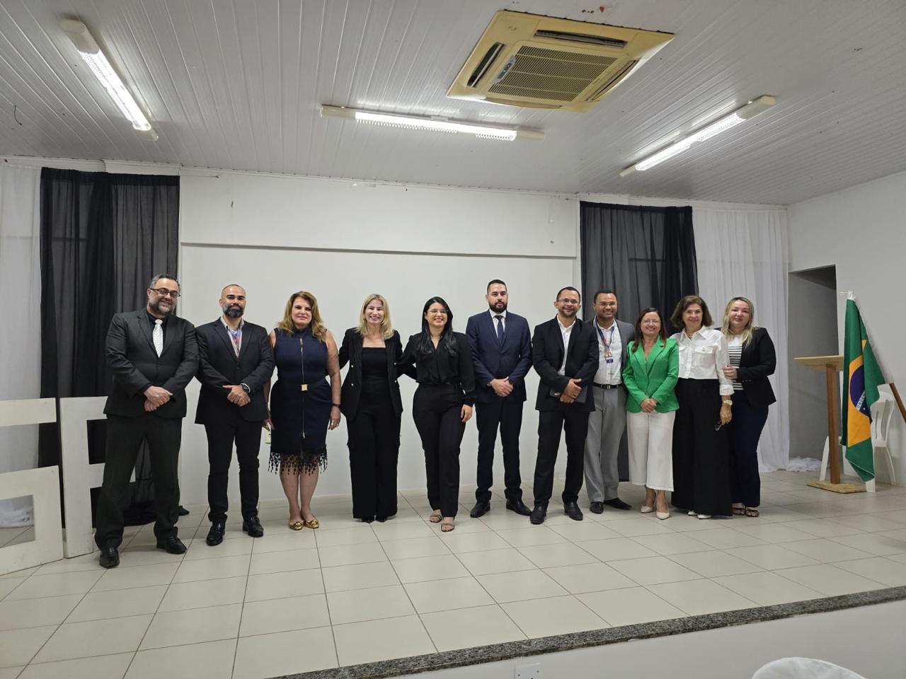 A imagem mostra sete pessoas em pé, lado a lado, posando para uma foto em um ambiente interno. Todos estão vestidos formalmente, com ternos e vestidos. O fundo é composto por cortinas pretas e brancas, e há letras grandes brancas no canto esquerdo da imagem. O chão é de azulejos claros e o teto tem luminárias fluorescentes.